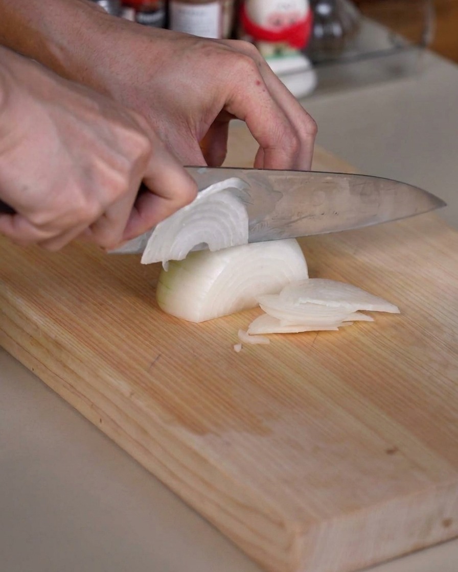 Un oignon en cours de découpe en fines lamelles sur une planche à découper en bois à laide dun couteau de cuisine bien aiguisé.