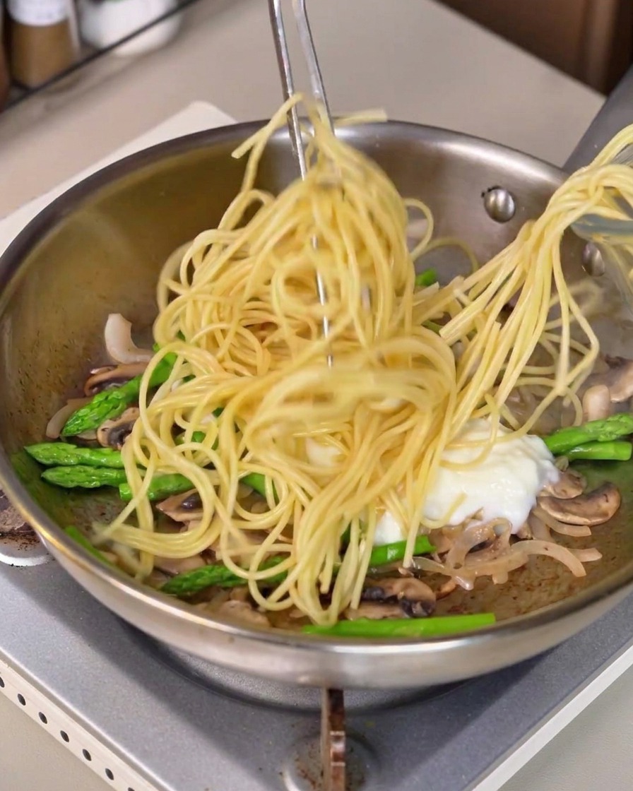 De longs spaghettis soulevés à laide de pinces et incorporés dans la poêle avec la sauce blanche et les légumes.