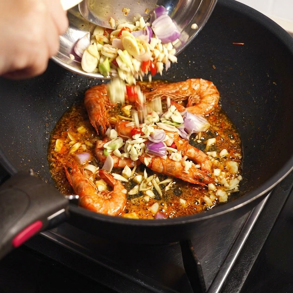 Aromates hachés et pâte Tom Yum ajoutés dans la poêle contenant les carapaces de crevettes sautées.