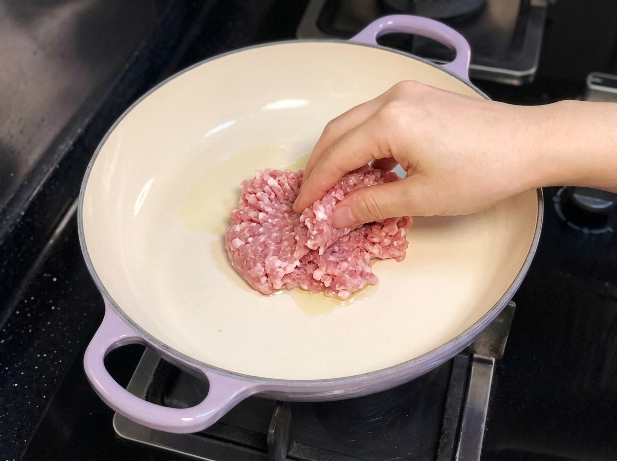 Une main déposant du porc haché dans une poêle à manche violet contenant de lhuile dolive sur une cuisinière.
