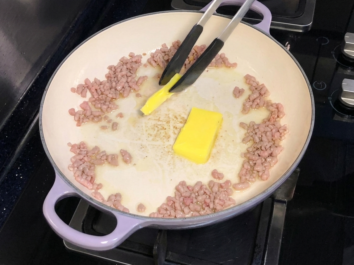 Du beurre ajouté et en train de fondre dans une poêle contenant du porc haché doré.
