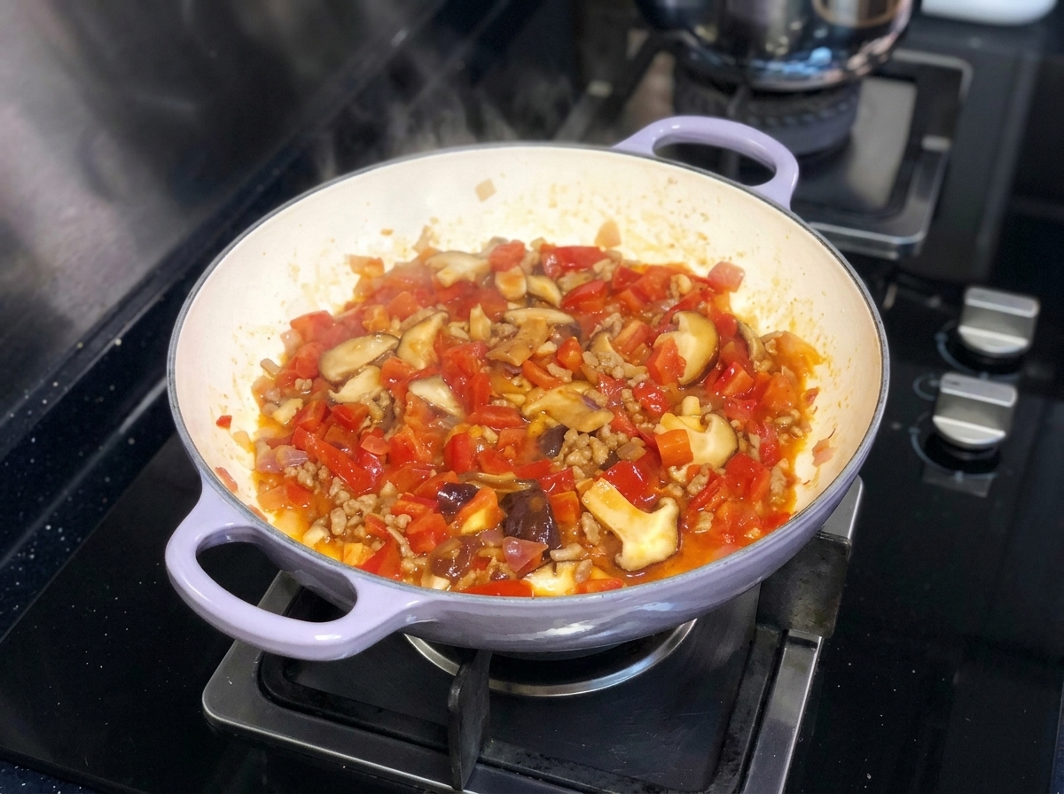 Un mélange vibrant de tomates hachées, doignons et de champignons shiitake mijotant ensemble dans une poêle sur une cuisinière.