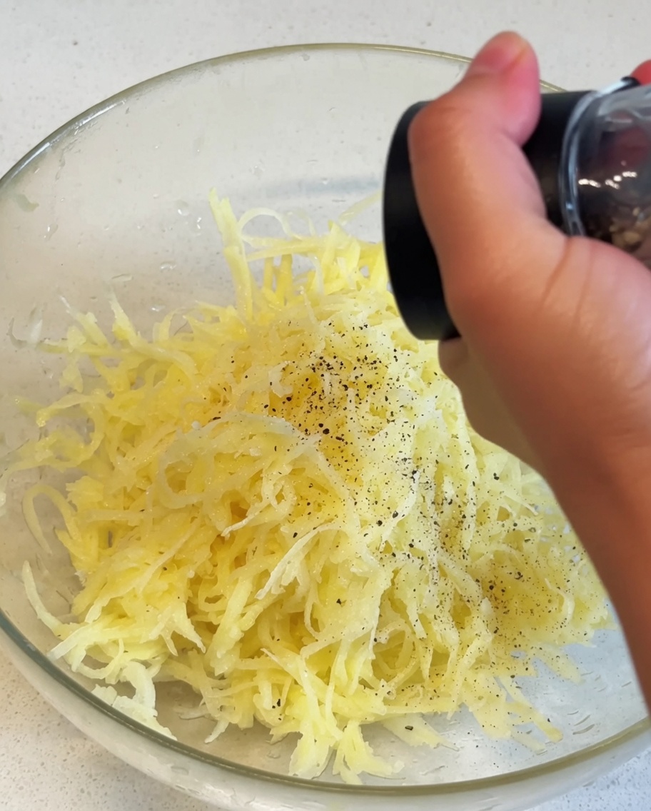 Pommes de terre jaunes râpées dans un bol en verre recevant un assaisonnement de poivre noir provenant dun moulin manuel.