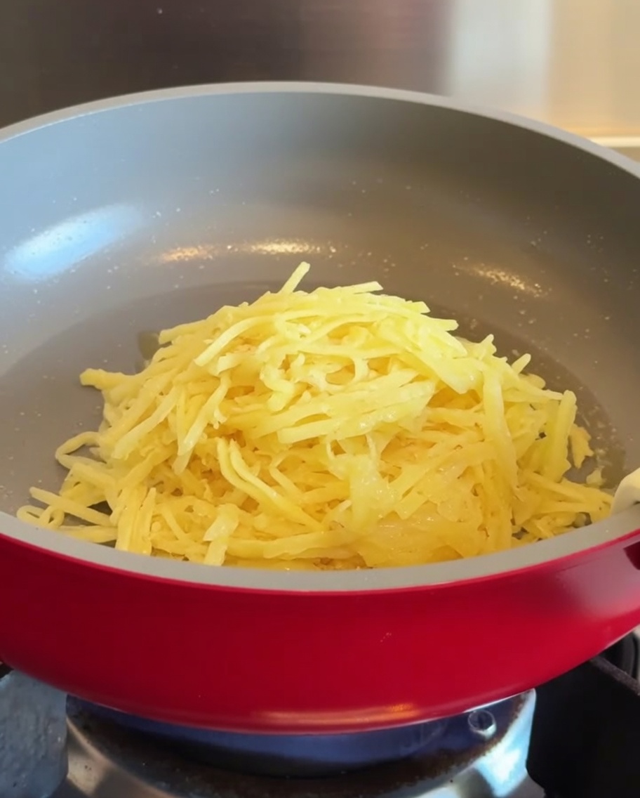 Un tas de pommes de terre râpées placé au centre dune poêle antiadhésive rouge chauffée.