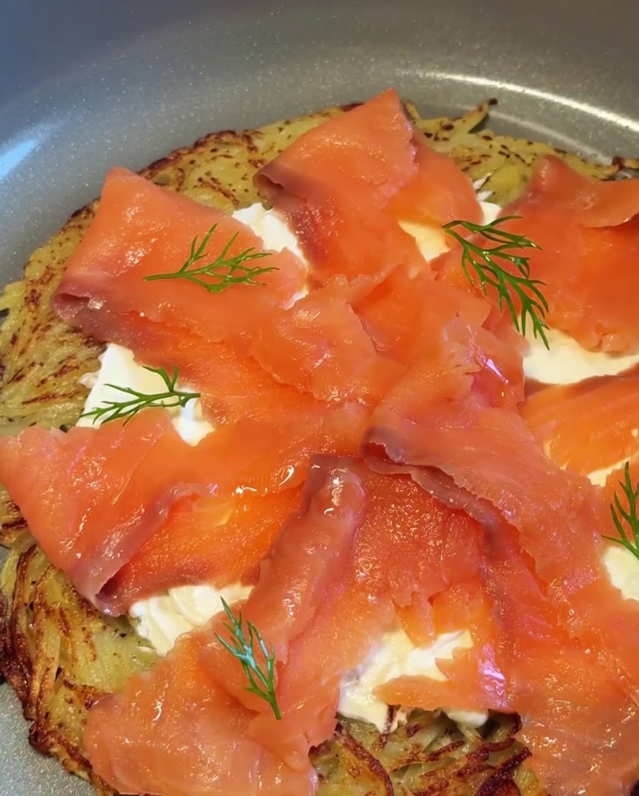 Une pizza aux pommes de terre et au saumon fumé terminée, garnie de brins daneth frais et de morceaux doignon rouge.