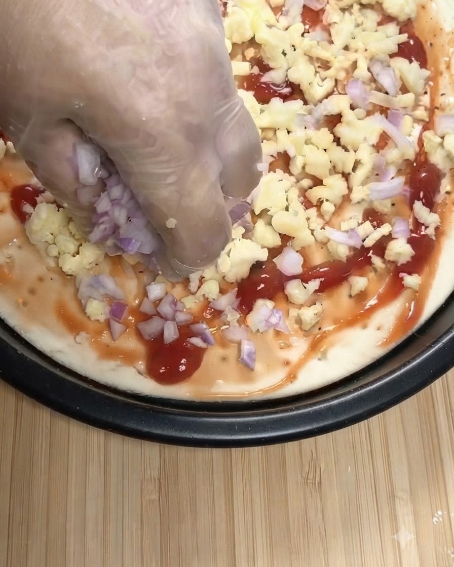 Une main portant un gant transparent saupoudrant des oignons rouges hachés et de la mozzarella râpée sur une base de pizza recouverte de sauce tomate.