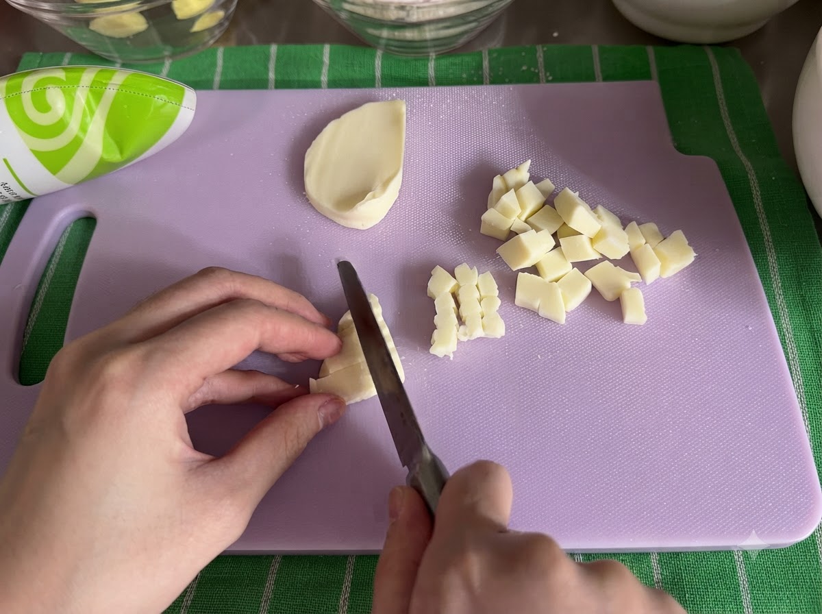 Un bloc de fromage mozzarella coupé en petits cubes avec un couteau sur une planche à découper.