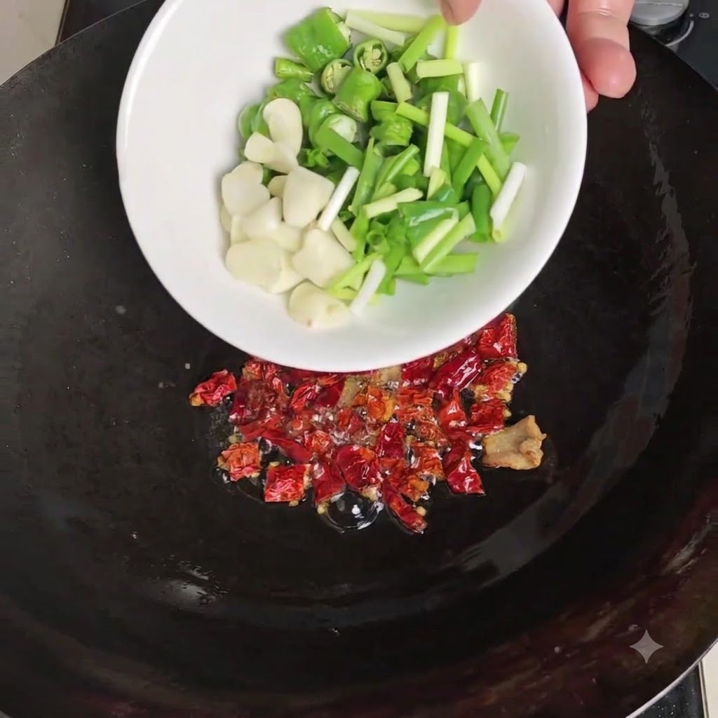 Un bol blanc versant des piments verts tranchés, de lail pelé et des ciboules sur les piments séchés torréfiés dans le wok.