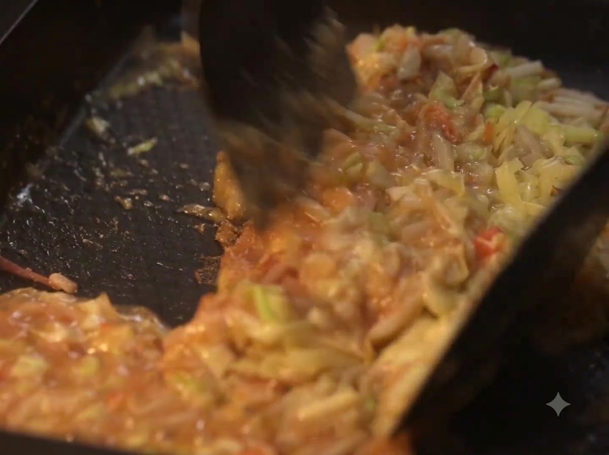 Des spatules mélangeant les légumes en cours de cuisson et la pâte liquide sur une plaque chauffante.