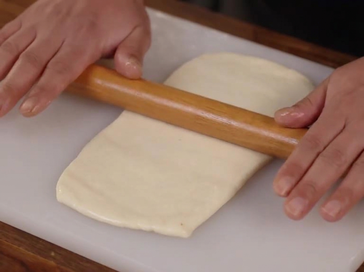 Des mains utilisant un rouleau à pâtisserie en bois pour aplatir un morceau de pâte lisse en un rectangle sur une planche à découper blanche.