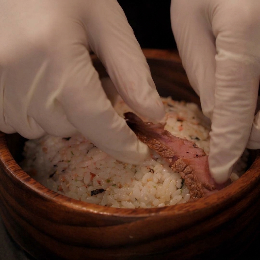 Des mains gantées plaçant des tranches de steak cuit sur un bol de riz assaisonné.
