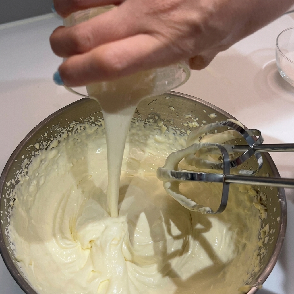 Une main versant de la crème supplémentaire dans un bol de crème fouettée et de beurre avec des batteurs électriques.