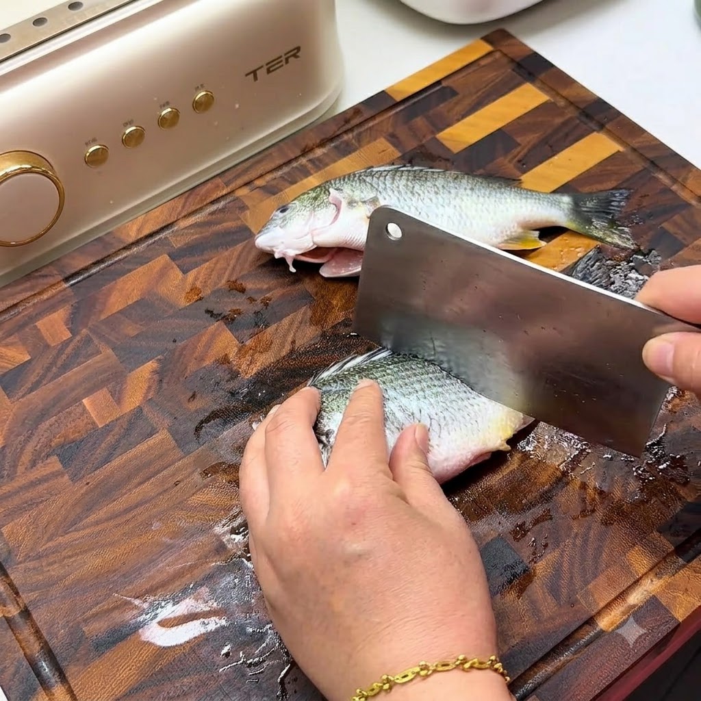 Une main utilisant un grand hachoir en métal pour faire des entailles en diagonale sur le flanc dun poisson frais sur une planche à découper en bois.