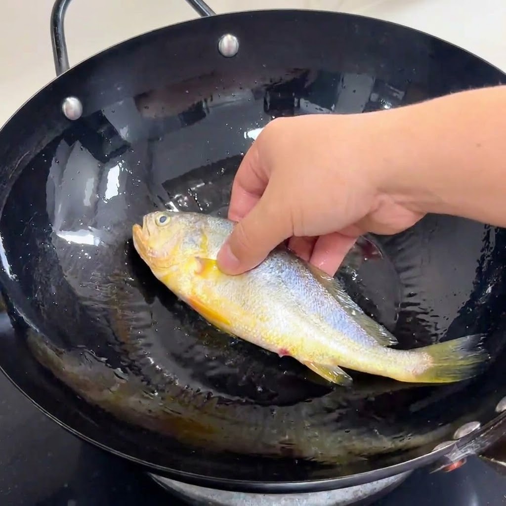Une main plaçant délicatement un poisson entier assaisonné dans un wok noir rempli dhuile de cuisson chaude.