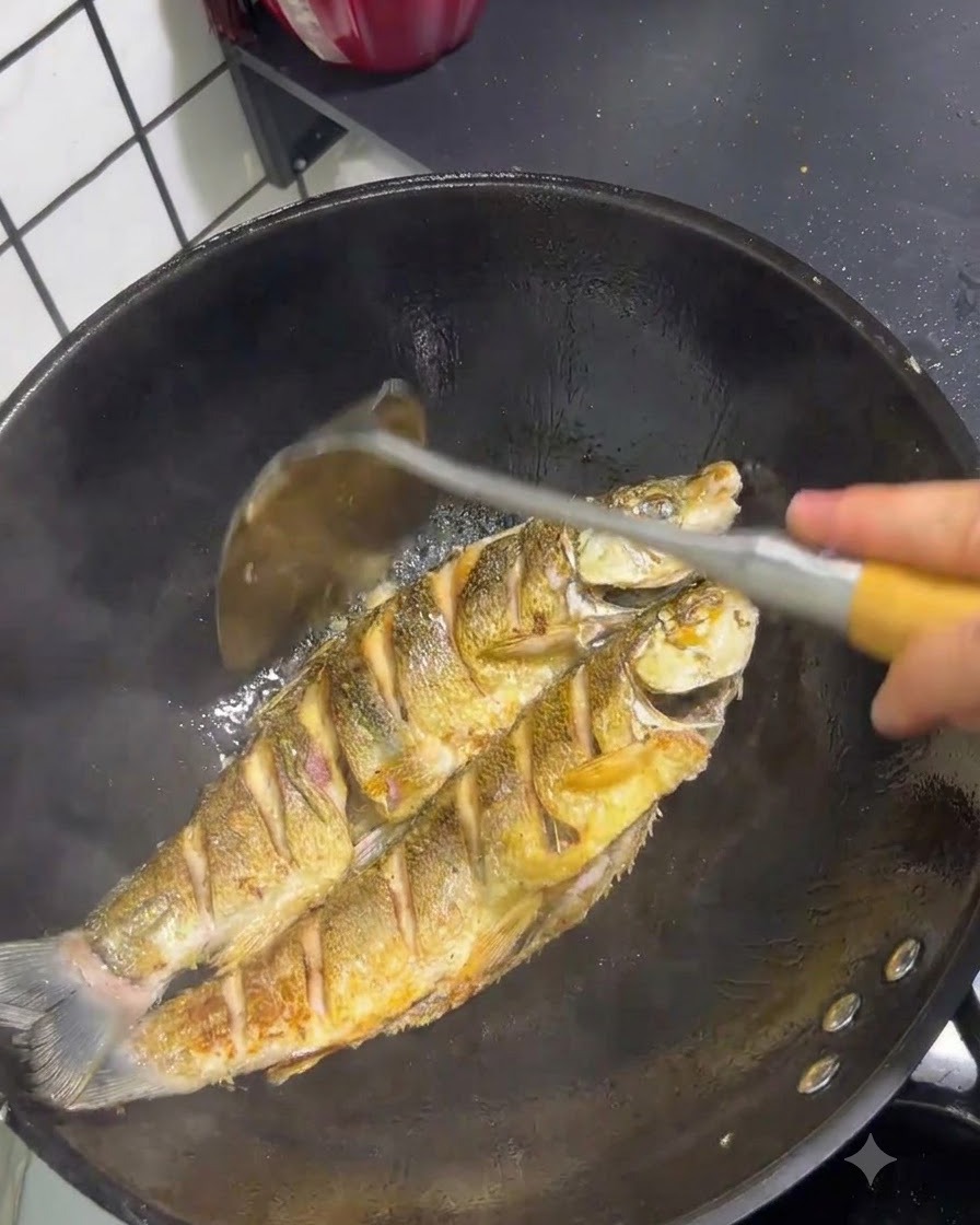 Deux poissons queue jaune en train de frire dans un wok avec une peau dorée manipulée par une spatule.