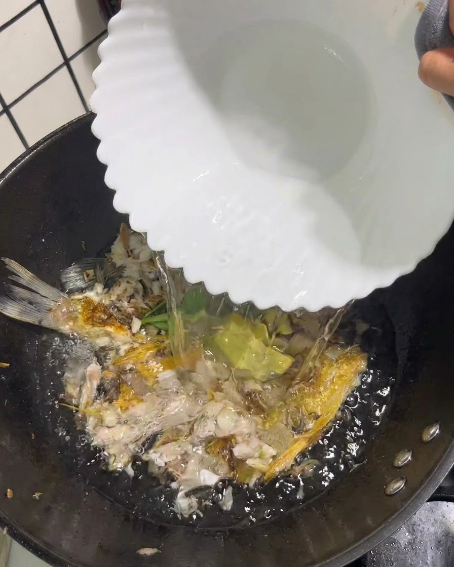 De leau bouillante claire versée dun bol blanc dans un wok contenant du poisson écrasé et des aromates.