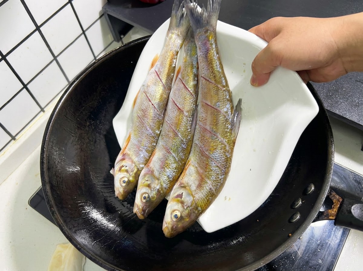 Trois poissons entiers et entaillés glissant dune assiette blanche vers un wok noir rempli dhuile chaude.