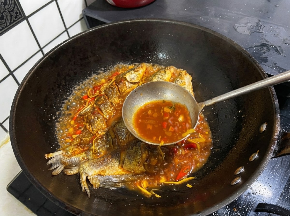 Une louche en métal puisant un bouillon sombre et savoureux dans une poêle contenant un poisson entier frit.