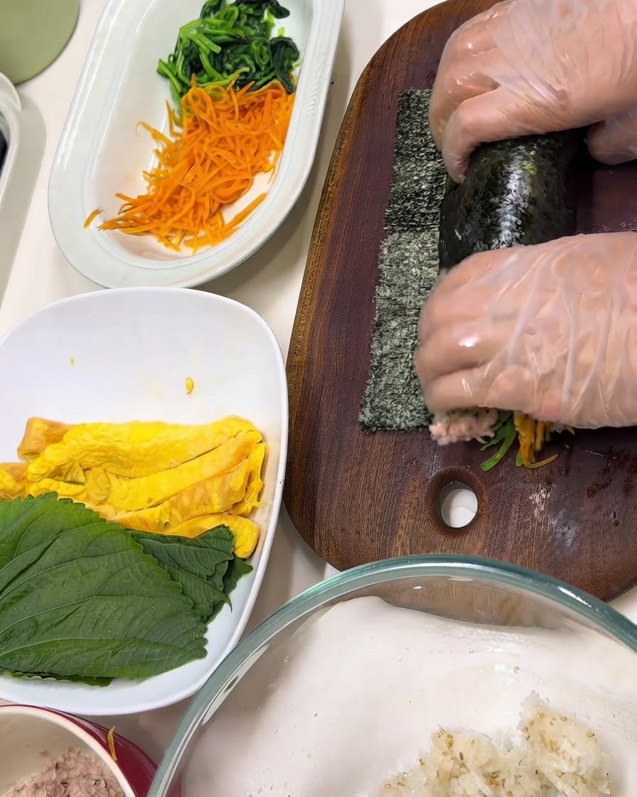Des mains portant des gants transparents enveloppant des ingrédients dans une feuille de nori sur une planche à découper en bois.