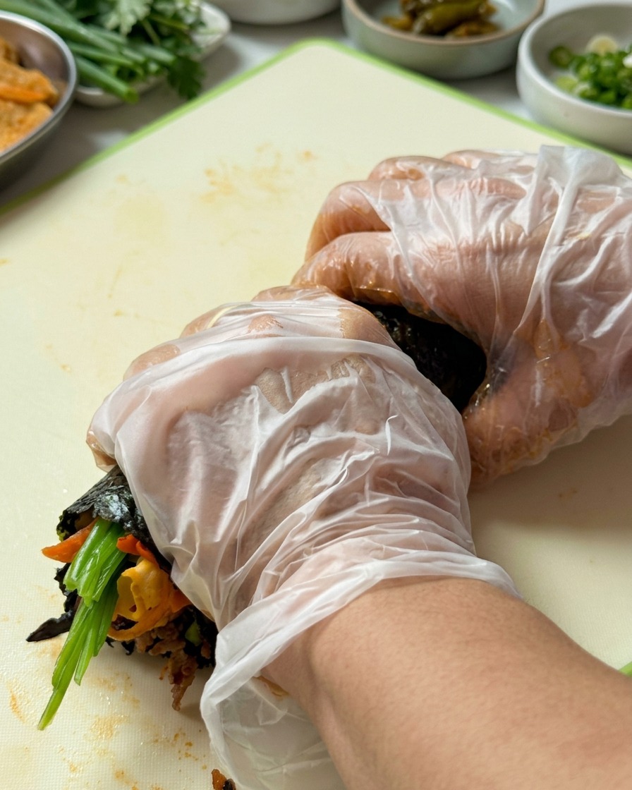 Mains gantées roulant fermement le gimbap du bord inférieur vers le haut.