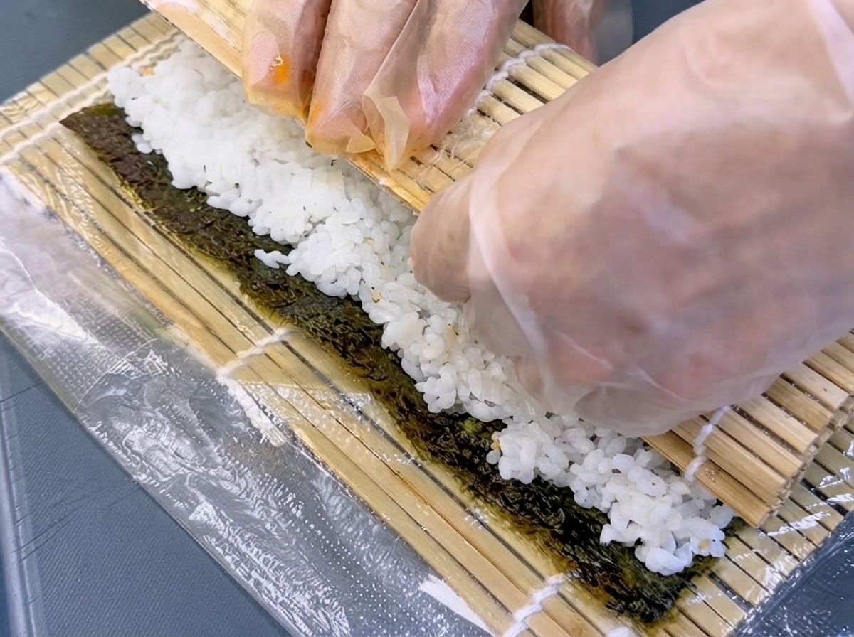 Des mains gantées soulevant une natte en bambou pour rouler fermement lalgue et sa garniture en un cylindre.