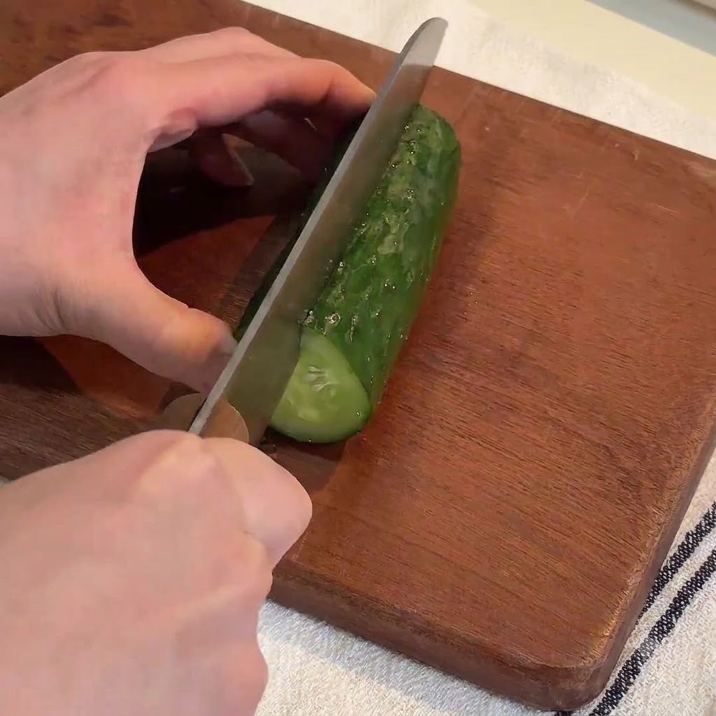 Mains utilisant un grand couteau pour couper un concombre vert frais en de longues lanières sur une planche à découper en bois.