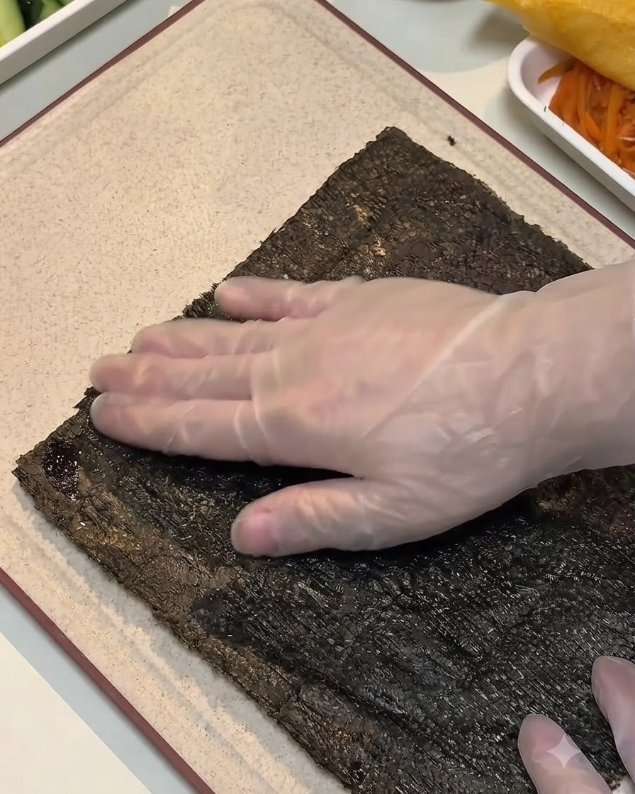 Une main gantée appuyant sur le bord dune feuille dalgue humidifiée sur une planche à découper.