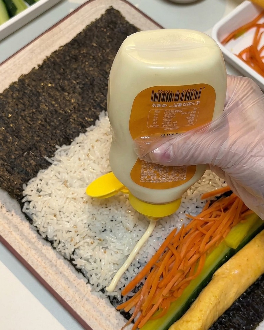 Une main gantée pressant des lignes de mayonnaise blanche dune bouteille à travers les garnitures disposées sur lalgue et le riz.