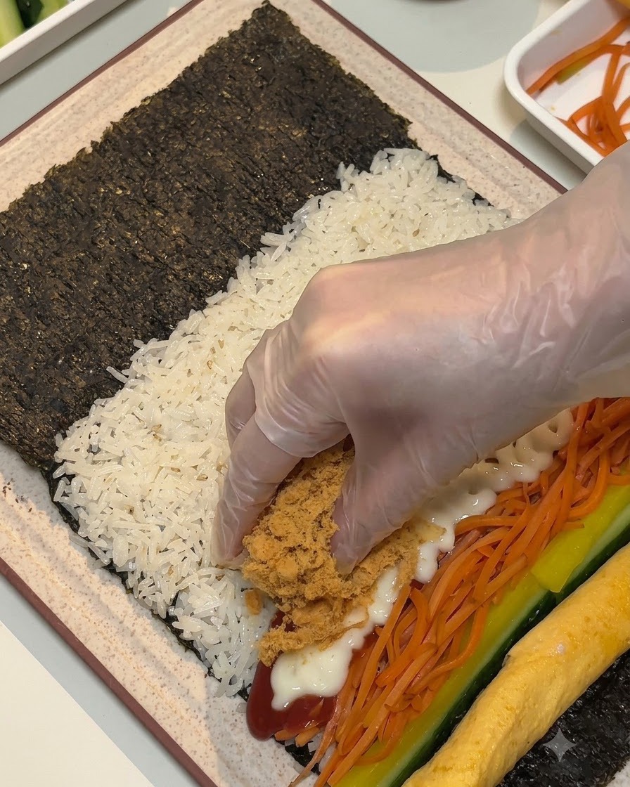 Une main gantée plaçant des flocons de viande bruns et duveteux et des lanières de viande sur les garnitures de la feuille dalgue.