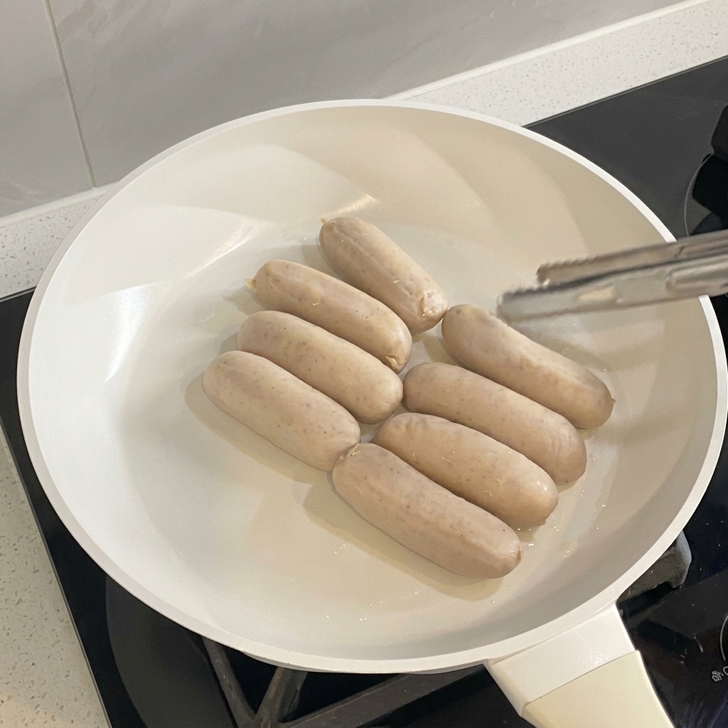 Saucisses en train de cuire et de dorer uniformément dans une poêle blanche.