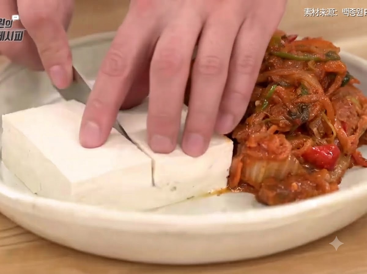 Une main utilisant un couteau pour trancher proprement du tofu blanchi sur une assiette à côté dun tas de kimchi sauté savoureux et de porc.