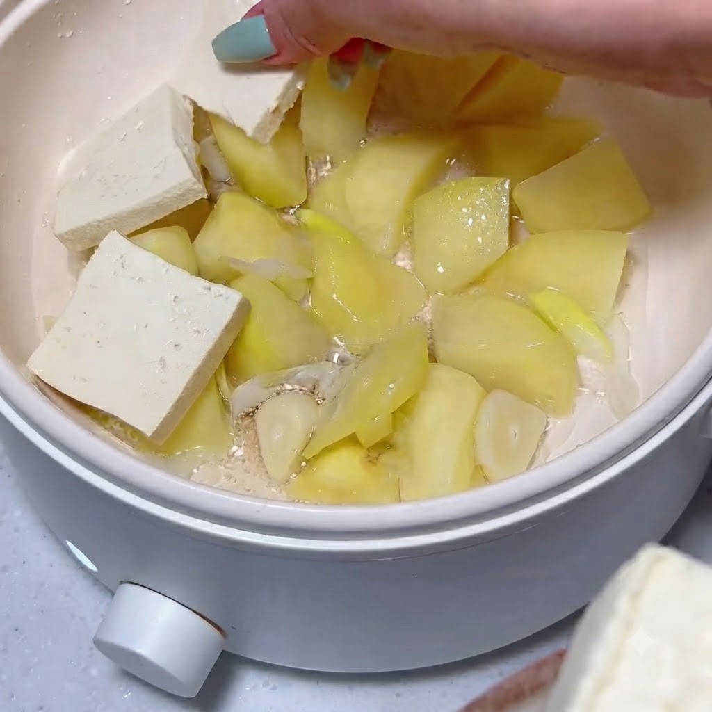 Des tranches de tofu soigneusement disposées sur une couche de morceaux de pommes de terre et dail cuits dans une marmite blanche.