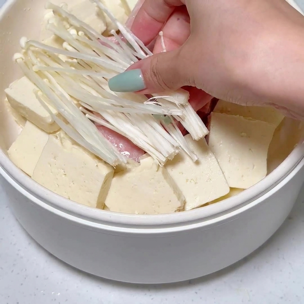 Des champignons enoki placés dans la marmite parmi les tranches de tofu et de luncheon meat.