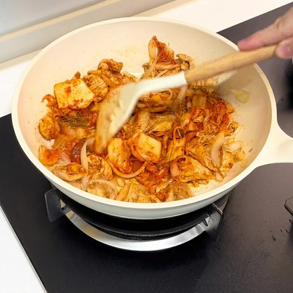 Du kimchi et du poulet sautés ensemble dans une poêle blanche à laide dune spatule.