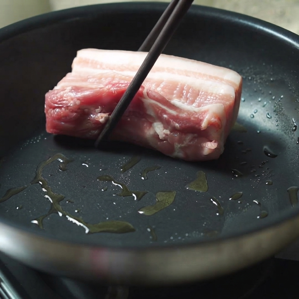 Un morceau épais de poitrine de porc en train dêtre placé dans une poêle chaude huilée à laide de baguettes.