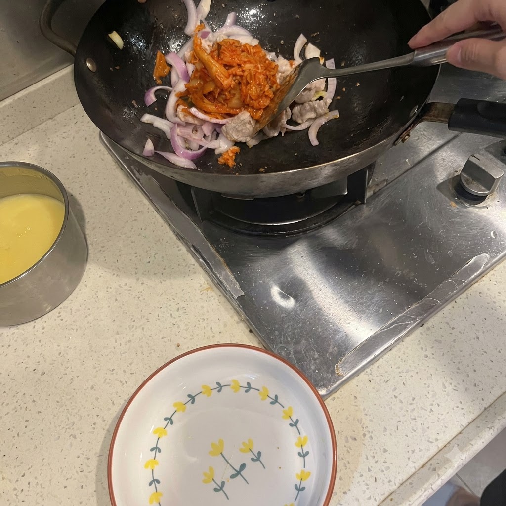 Une spatule ajoutant du kimchi rouge vif dans un wok contenant de la poitrine de porc et des oignons rouges sautés.