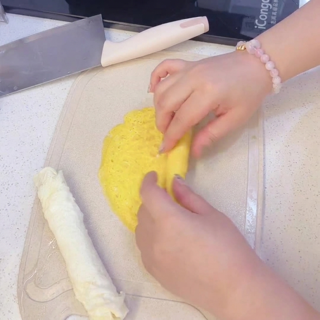 Mains roulant une fine crêpe dœuf jaune sur une planche à découper, à côté dune crêpe dœuf blanche déjà roulée.
