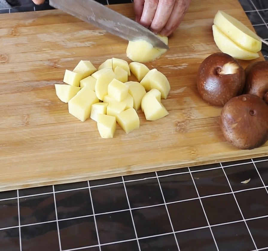 Mains coupant des pommes de terre jaunes pelées en petits cubes sur une planche à découper en bambou.