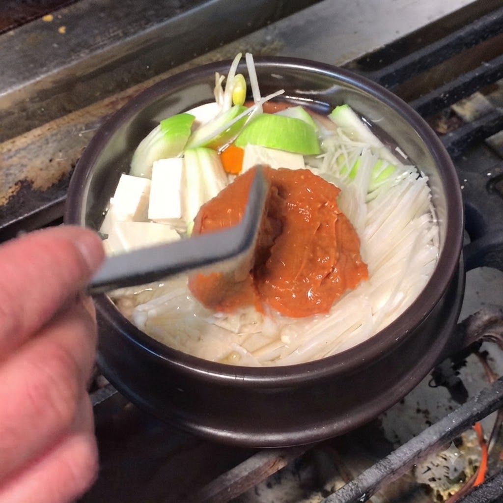 Une spatule en silicone ajoutant de la pâte de soja brune riche et de la pâte de piment rouge dans la casserole.