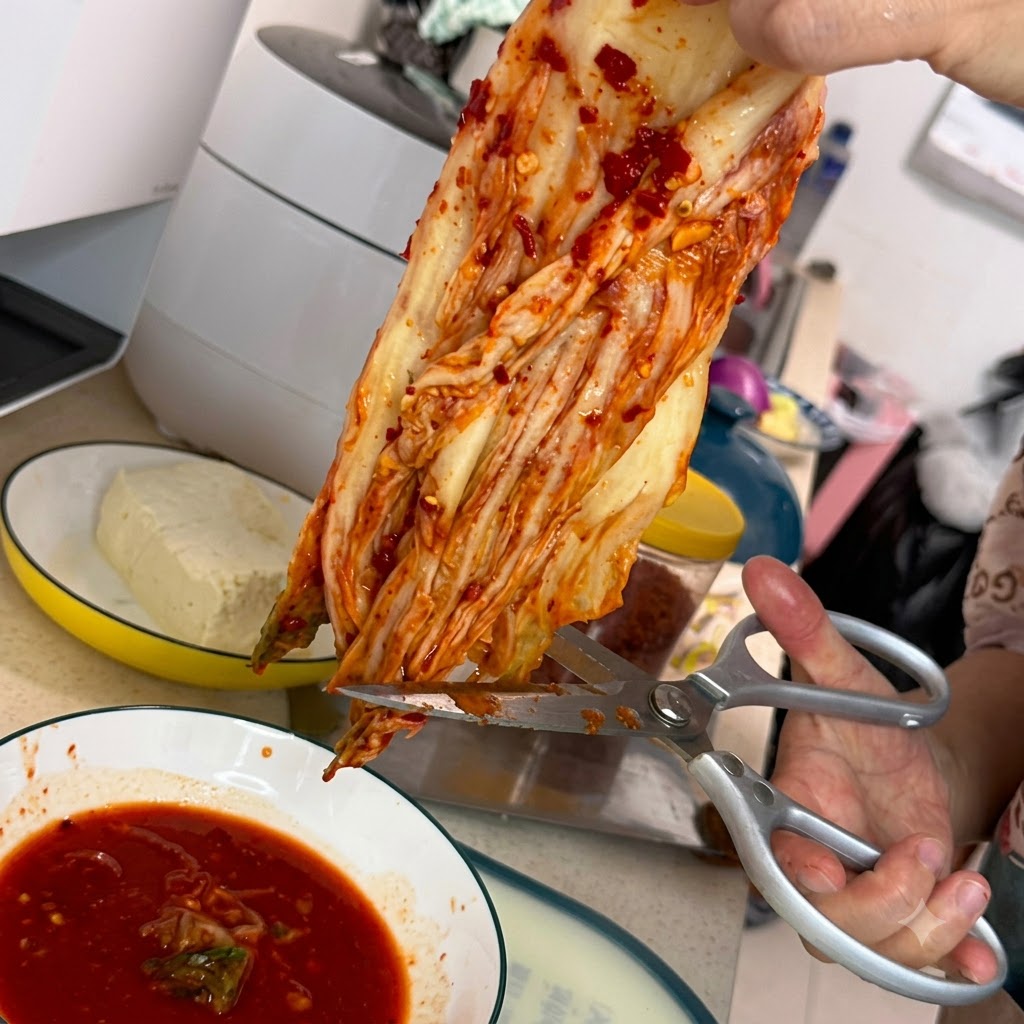 Une paire de ciseaux de cuisine argentés coupant un morceau de kimchi rouge au-dessus dun bol blanc rempli de sauce rouge épicée.