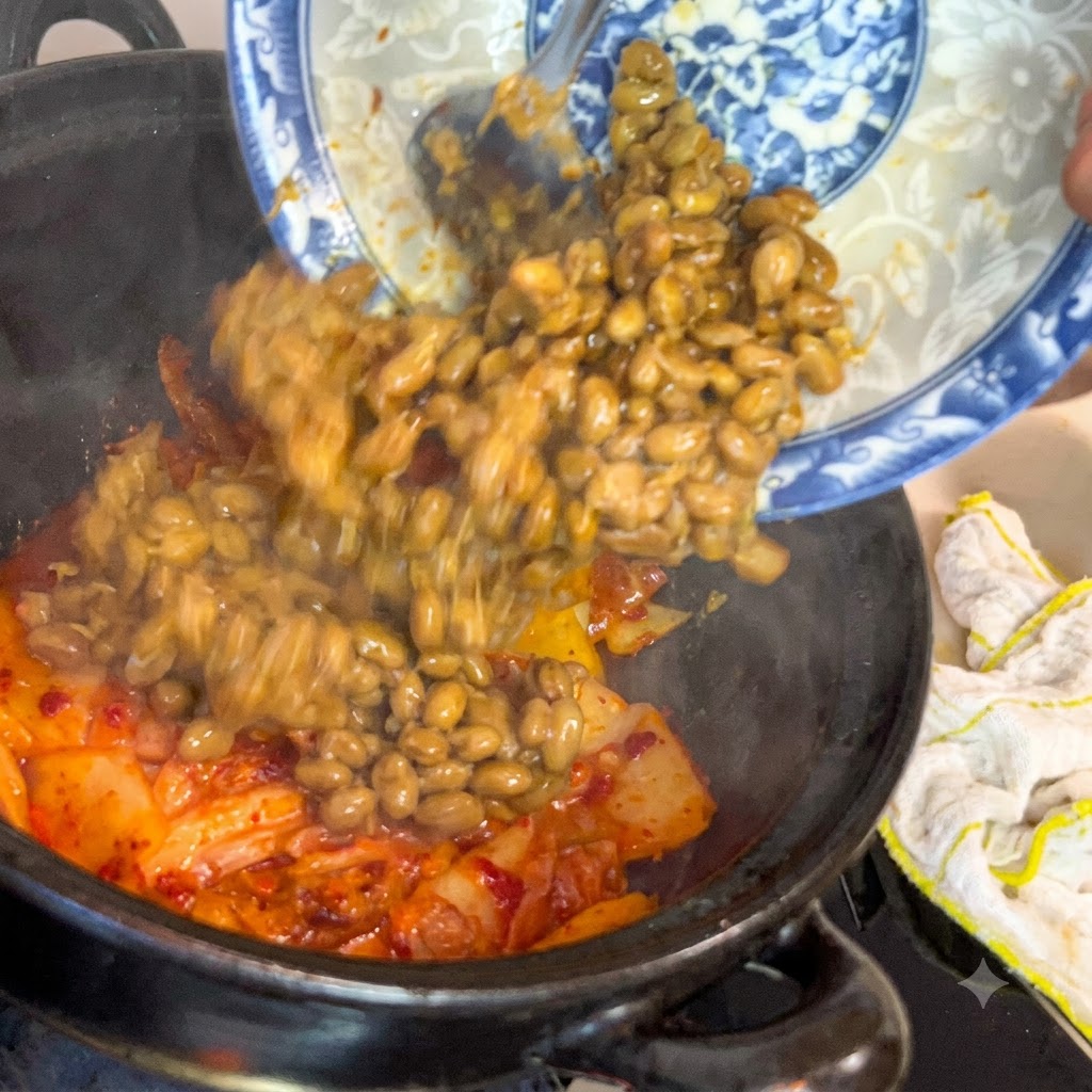 Des graines de soja fermentées glissant dune assiette décorative bleue et blanche dans une casserole sombre remplie de kimchi grésillant.