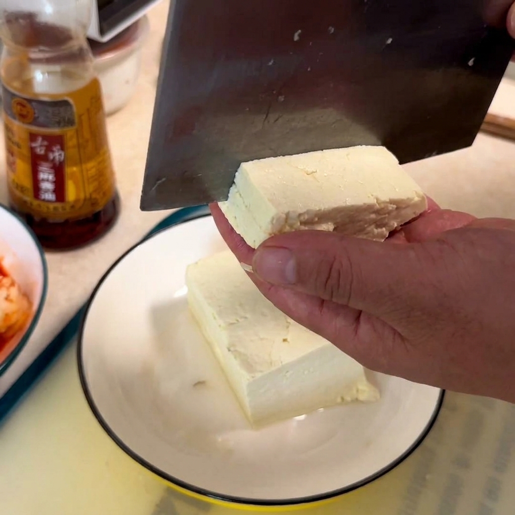 Une main tenant un bloc de tofu blanc tandis quun hachoir le coupe en cubes sur une assiette blanche.