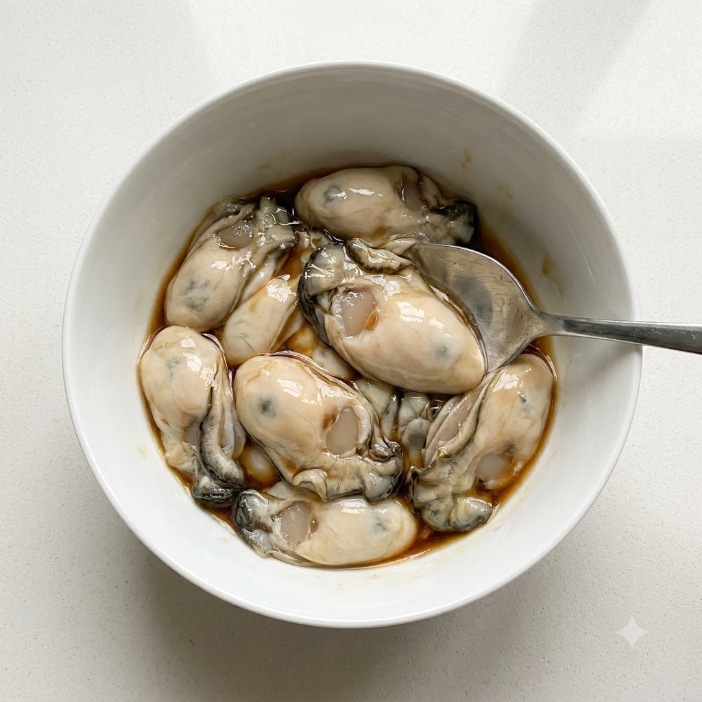 Des huîtres nettoyées dans un bol blanc assaisonnées avec de la sauce nuoc-mâm et du poivre blanc en poudre à laide dune cuillère.