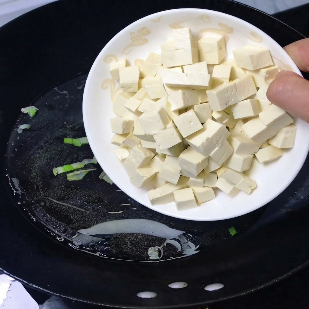 Une assiette de tofu blanc en cubes versée dans un wok noir rempli deau bouillante.