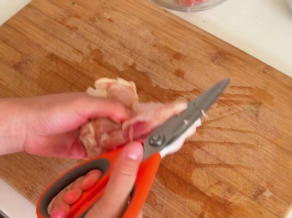 Une paire de ciseaux de cuisine découpant la viande autour de los dune cuisse de poulet crue sur une planche en bois.