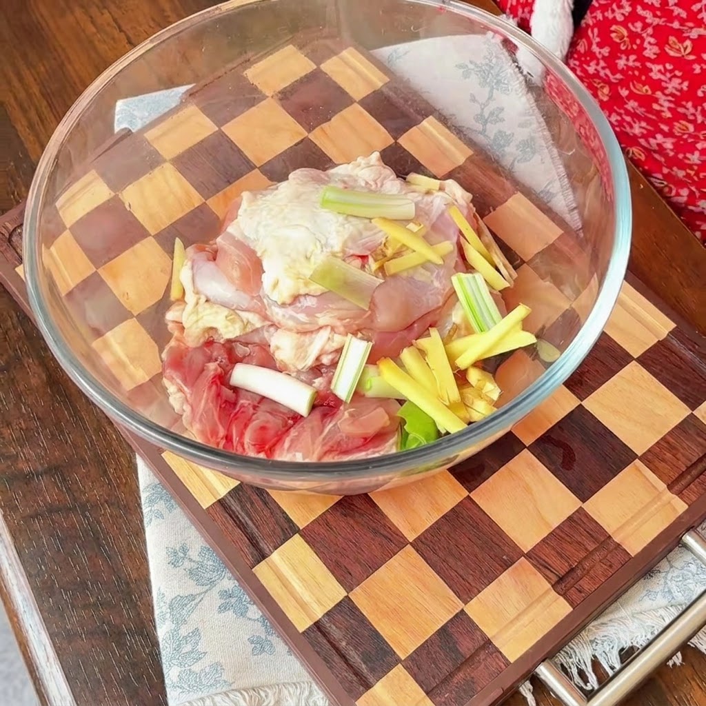 Morceaux de poulet crus dans un bol en verre recouverts de tranches doignons rouges, de cébettes et de bâtonnets de gingembre.
