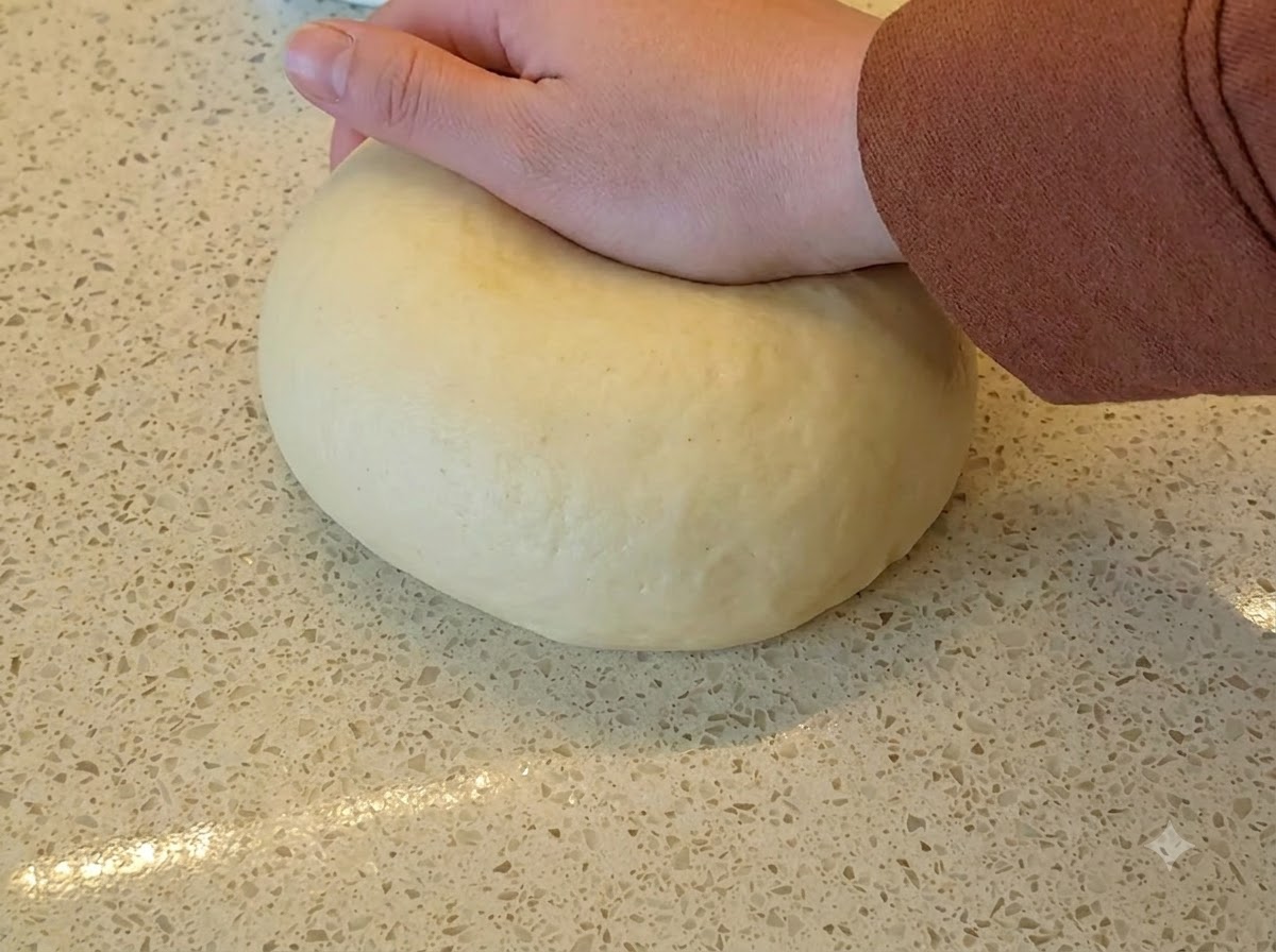 Une main appuyant sur une grande boule lisse de pâte à pain jaune sur un plan de travail moucheté.