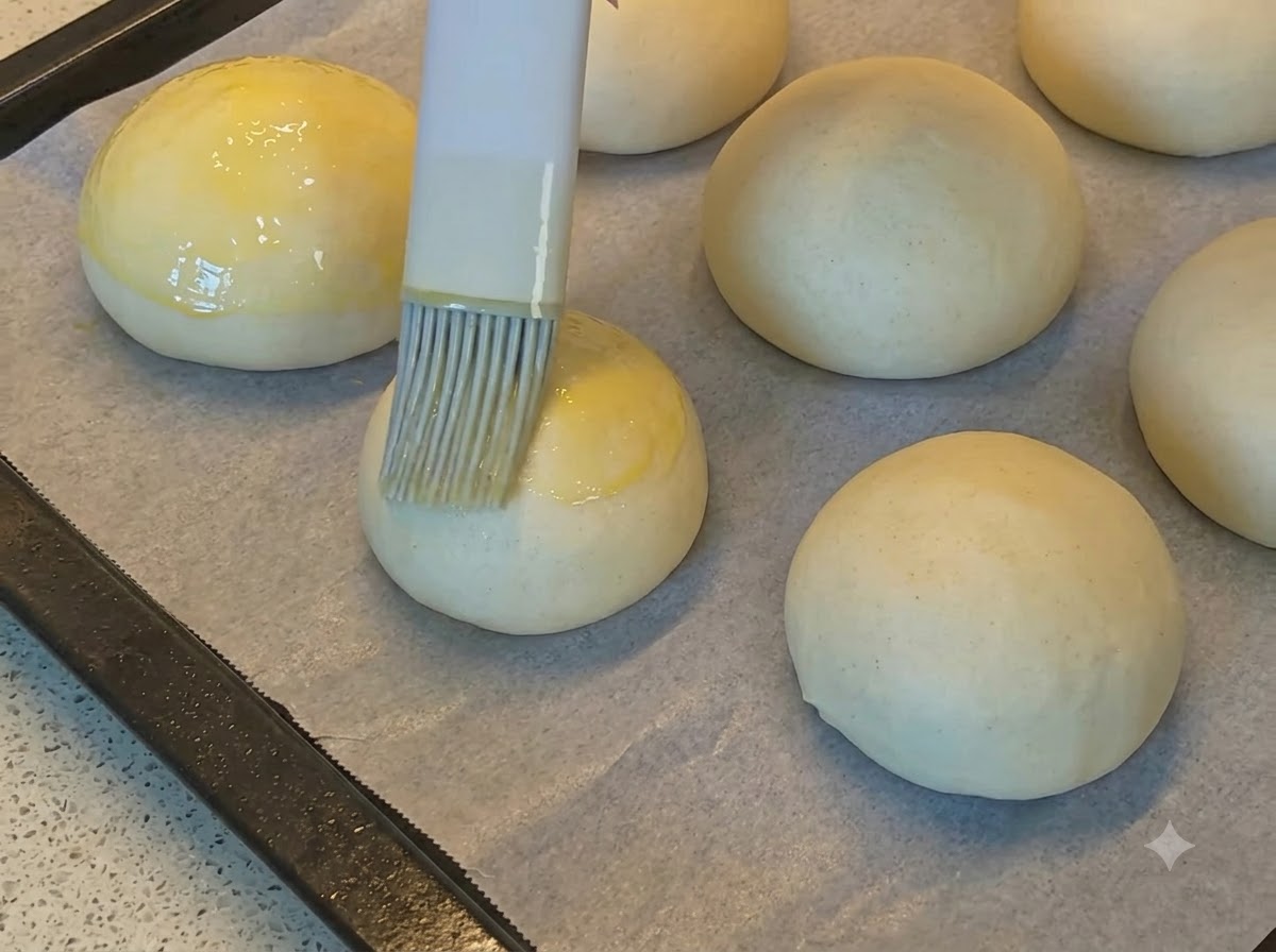 Un pinceau de cuisine appliquant une dorure à lœuf dorée sur des boules de pâte à pain rondes et lisses disposées sur du papier sulfurisé.