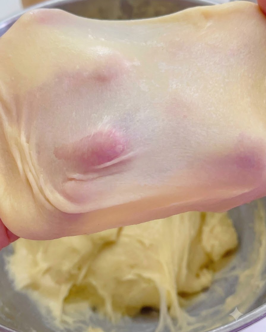 Pâte à brioche étirée montrant une fine membrane de type fenêtre translucide tenue par les doigts.