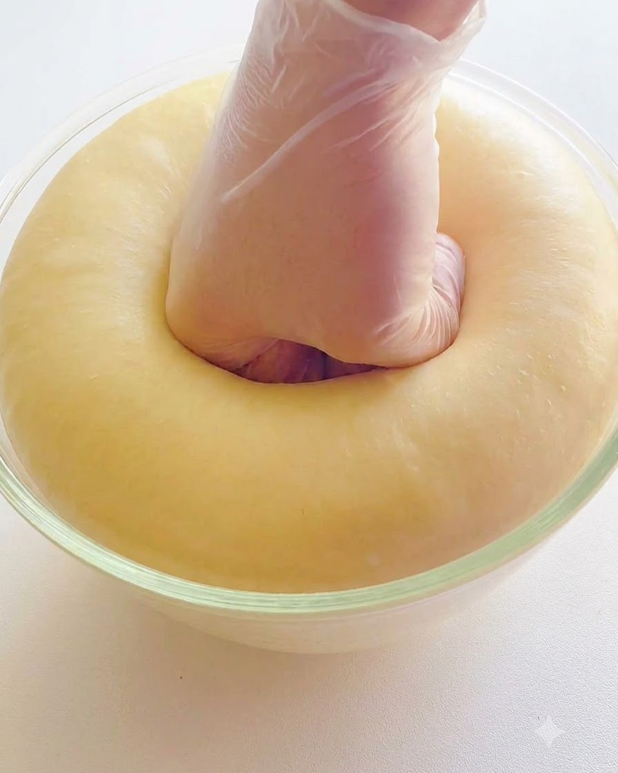 Main gantée de plastique enfonçant le centre dune pâte bien levée et bouffie dans un bol en verre.